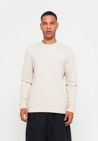 Sudadera de cuello redondo en color beige claro, fabricada con un tejido suave. Presenta mangas largas y un dobladillo ajustado y acanalado. Pequeño logo en el lado izquierdo del pecho.