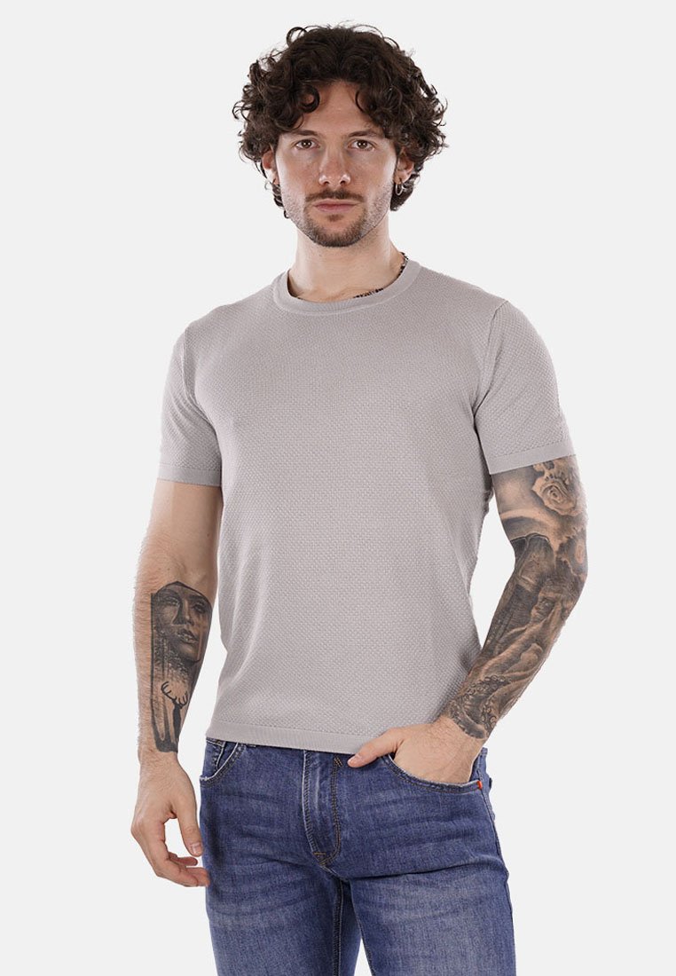 Camiseta gris de manga corta con tejido texturizado y escote redondo, combinada con jeans de mezclilla azul. Presenta tatuajes visibles en los brazos.