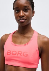 Γυναίκα που φοράει φωτεινό ροζ αθλητικό σουτιέν με τη λέξη "BORG" τυπωμένη με έντονα γράμματα μπροστά, πάνω σε απλό ανοιχτό φόντο.