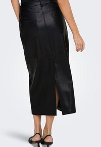 Femme portant une jupe midi en cuir noir avec des poches arrière et une fente centrale, associée à des sandales à talons noires.