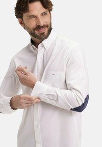 Homme avec une barbe qui attache le poignet d'une chemise blanche à boutons avec des coudières bleu marine et un petit détail brodé rose sur la poche.