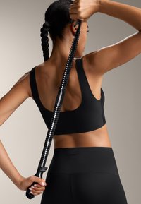 Czarny stanik sportowy z szerokimi ramiączkami, o gładkiej teksturze. Połączony z czarnymi legginsami, trzymający teksturowaną taśmę oporową w jednej ręce.