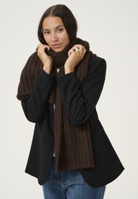 Écharpe marron à côtes en laine texturée, portée autour du cou. Associée à un blazer noir et un jean en denim, avec une coupe décontractée.