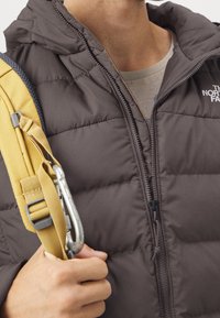 Bruine gewatteerde bodywarmer met capuchon, voorzien van een ritssluiting aan de voorkant en doorgestikte patronen. In de linkerhand wordt een gele riem en een karabijnhaak vastgehouden.