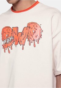 Crewneck t-skjorte i lys beige med rød hals. Har et smeltende, blankt oransje grafikk med bokstavene "CWP" i drypp-effekt.