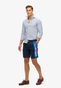 Chemise boutonnée à rayures bleu clair, short de bain bleu marine avec imprimé de logo rouge et bleu, chaussures bateau marron, montre au poignet gauche.