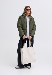 Olivgrüne Steppjacke mit Kapuze, grauer Kapuzenpullover darunter, schwarze weite Hose und beige Tragetasche. Schwarze Sonnenbrille, weiße Schuhe.