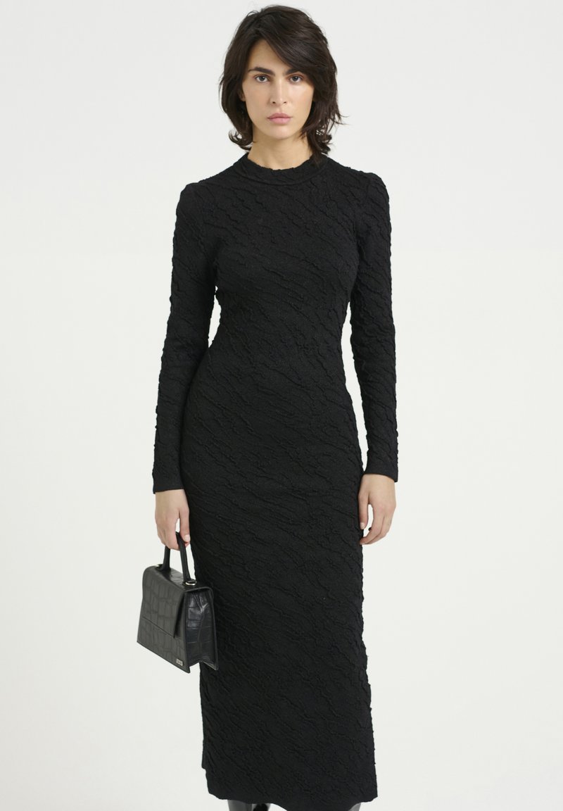 Abito nero a maniche lunghe realizzato in tessuto testurizzato, con silhouette aderente e scollatura rotonda. La modella tiene una piccola borsa nera con un design strutturato.