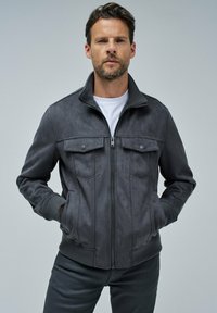 Chaqueta de ante gris con cierre de cremallera en la parte delantera, puños y dobladillo acanalados. Presenta dos bolsillos en el pecho con botones de presión y un diseño de cuello alto.