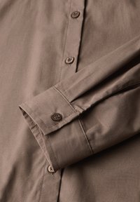 Camicia marrone a maniche lunghe con polsini abbottonati e bottoni visibili sulla patta anteriore, realizzata in tessuto liscio.