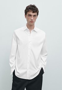 Camicetta bianca a maniche lunghe con colletto classico, realizzata in un tessuto liscio, caratterizzata da una vestibilità rilassata e una leggera lucentezza. Pantaloni neri.