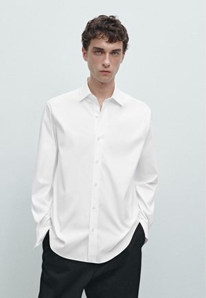 Chemise blanche à manches longues avec un col classique, en tissu lisse, présentant une coupe décontractée et un éclat subtil. Pantalons noirs.