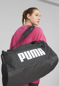 Black duffel bag with a large white "PUMA" logo, features a textured exterior, adjustable strap, and mesh accents on the side.
