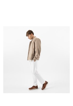 Junger Mann mit Händen in den Taschen, trägt eine beige Jacke, weiße Hose und braune Sneakers vor weißem Hintergrund.