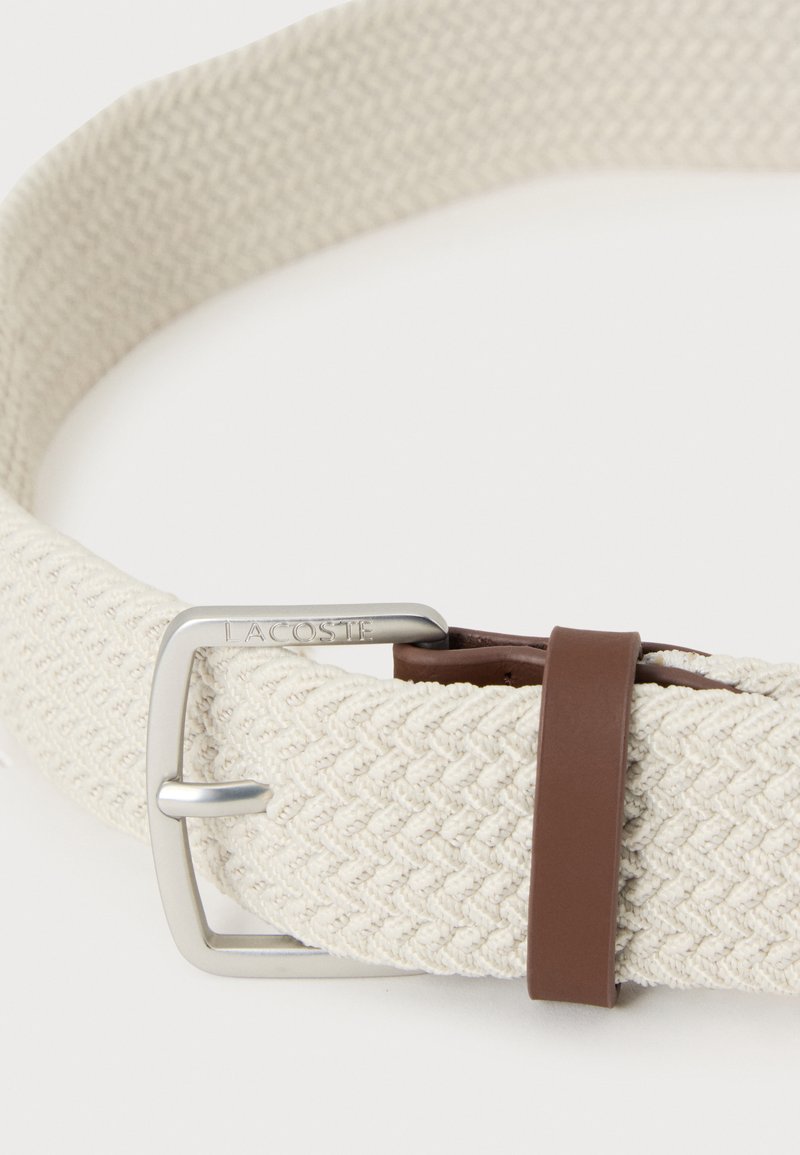 Off-white woven fabric belt with silver metal buckle engraved "Lacoste" and a brown leather loop near the buckle.