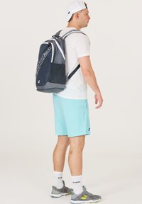 Mochila gris y negra con acentos blancos y un bolsillo de malla, llevada en la espalda de una persona que viste una camiseta blanca y pantalones cortos azul claro.