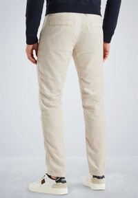 Beige katoenen broek met een slim fit, voorzien van twee achterzakken en een gladde textuur, gecombineerd met witte sneakers en een donker sweatshirt.