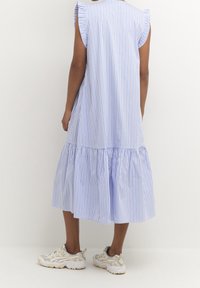 Baksidan av en person som bär en lös, ärmlös, ljusblå och vitrandig midi-klänning med volangkant och beige sneakers.