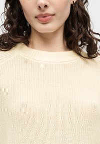 Woman with curly brown hair wearing small gold earrings and a cream-colored knitted sweater with a ribbed crew neckline.