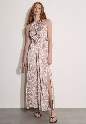 Frau in ärmellosem Halterneck-Maxikleid mit pinkem botanischem Muster und Seitenschlitz, trägt eine Halskette mit Anhänger und braune Sandalen.