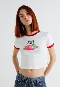 Lyhythihainen crop-top, valkoista kangasta, punaisilla pikeillä, jossa on mansikkagraafinen kuva vihreillä lehdillä ja valkoisilla kukilla.