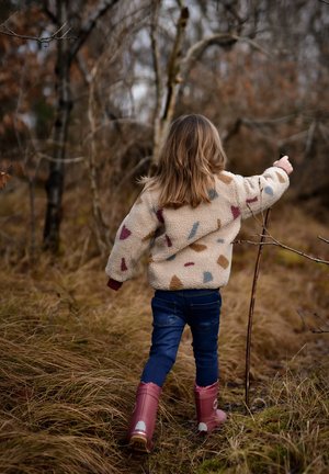 Dítě v krémové fleecové bundě s barevnými tvary, tmavými džínami a růžovými gumovými botami, procházející se vysokou trávou v zalesněné oblasti.