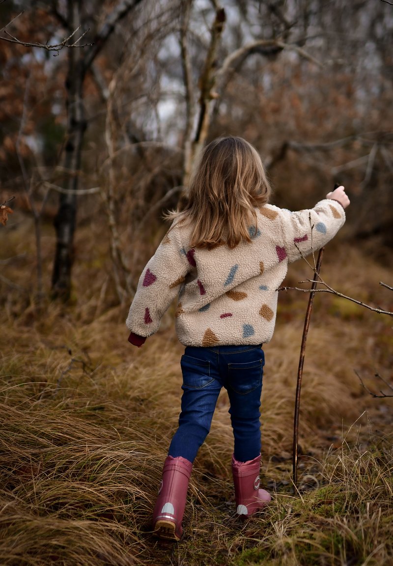 Gyermek krémszínű polárkabátban, színes mintákkal, sötét farmerral és rózsaszín gumicsizmával, magas fű között sétál egy erdős területen.