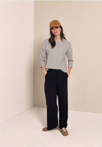 Gestreifte Langarmbluse in Weiß und Marineblau, kombiniert mit weit geschnittenen Marineblauen Hosen und einer braunen Kappe. Braune Sneaker mit weißen Schnürsenkeln runden das Outfit ab.
