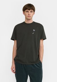 Camiseta de algodón verde oscuro con escote redondo y mangas cortas. Presenta un pequeño gráfico gris cerca del pecho. Corte relajado.