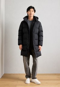 Schwarze Daunenjacke mit Kapuze, gestepptem Design und Druckknöpfen, getragen über einem grauen Pullover. Kombiniert mit grauen Jeans und weißen Sneakers.