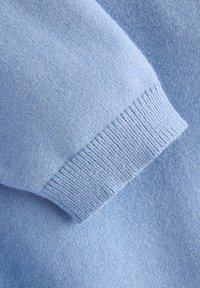 Tissu de pull en tricot bleu clair avec une texture lisse, présentant un poignet côtelé et de subtiles variations de teinte et de tissage.