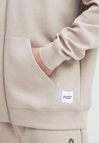 Veste à capuche zippée beige clair avec une texture douce, dotée d'une poche kangourou et de poignets côtelés, marquée d'une étiquette "North Bend".