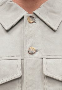 Giacca in suede grigio chiaro con colletto pieghevole, dotata di due bottoni. Dettagli di cucitura visibili sulle tasche. Texture morbida e leggera lucentezza.