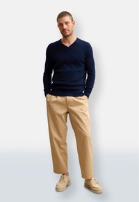 Marinblå v-halströja, beiga veckade byxor och ljusbruna skor. Mjuk textur, avslappnad passform. Neutral bakgrund som förstärker outfitens synlighet.