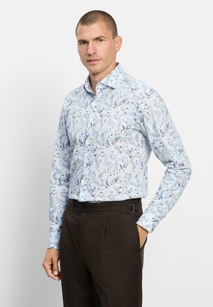 Mann trägt ein hellblaues, paisley-mustertes Langarmhemd und dunkelbraune Hose, steht mit einer Hand in der Tasche.