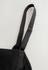 Bretelle de soutien-gorge en tissu noir réglable avec un bord bouclé et un curseur en plastique sur un fond clair.
