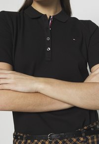 Svart polo-shirt i en texturerad stickning med krage och tre-knappsknop. Har en subtil logotyp och kontrasterande randigt innerknäpp.