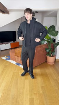 Giacca nera con zip sopra una maglietta nera, abbinata a pantaloni neri e scarpe nere. La giacca ha un colletto alto e una vestibilità rilassata.