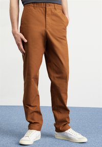 Brown cotton trousers with a tailored fit, featuring side pockets and a button fly. Paired with white sneakers on a blue carpet background.