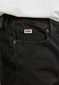 Jeans di denim neri con un primo piano della tasca posteriore. Presentano un'etichetta con il logo "GUESS" rosso e bianco e dettagli in metallo argentato.