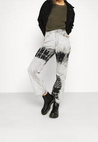 Hoch taillierte, gerade geschnittene Jeans in Hellgrau mit schwarzem Batikmuster und kontrastierenden Nähten, kombiniert mit schwarzen Knöchelboots und einem dunklen Oberteil.