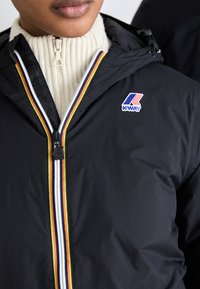 Schwarze K-Way-Jacke mit mehrfarbigen Reißverschlussdetails und Logo, getragen über einem cremefarbenen Ripp-Strickpullover. Sanftes, leichtes Material.
