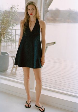 Młoda kobieta z długimi włosami, ubrana w czarną sukienkę bez rękawów i czarne sandały, stoi w pomieszczeniu przy dużym oknie z widokiem na taras.