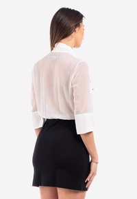 Hvid gennemsigtig bluse med krave og oprullede ærmer, parret med en sort tætsiddende nederdel. Stoffet er let og let gennemsigtigt.