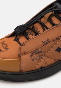 Brun sneakers med en struktureret overflade og et logo-mønster, sorte snørebånd og en kontrasterende gummisål. Sømme med syede detaljer.