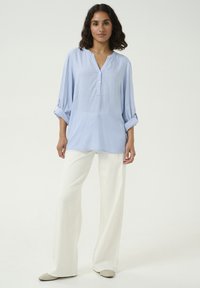 Lichtblauwe, langmouwige blouse met een V-hals en knoopsluiting, gecombineerd met witte wijde broek en neutrale instappers.