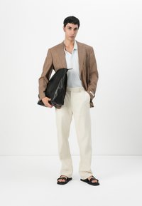 Braun karierter Blazer über einem hellen Polo, cremefarbene Hose, schwarze Sandalen, hält eine schwarze Ledertasche. Einfaches Design, neutrale Farben.
