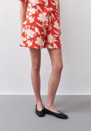 Femeie purtând pantaloni scurți roșii cu flori și cămașă asortată, combinată cu pantofi comozi negri cu vârf pătrat, stând pe podea gri.