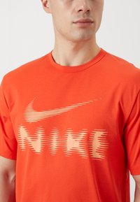 Punane puuvillane t-särk, millel on rinnal suur beež Nike'i logo kiirustriipude kujundusega. Lühikesed varrukad ja ümmargune kaelus.