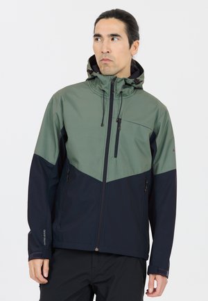 Homme portant une veste à capuche vert et noir avec des poches zippées, regardant sur le côté devant un fond blanc uni.